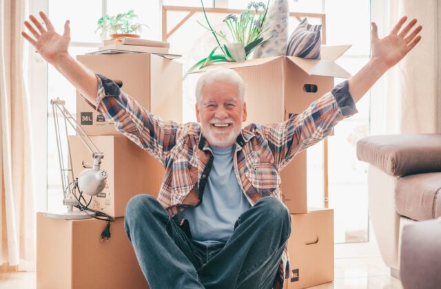 A happy senior man sitting on the floor in front of packed moving boxes.