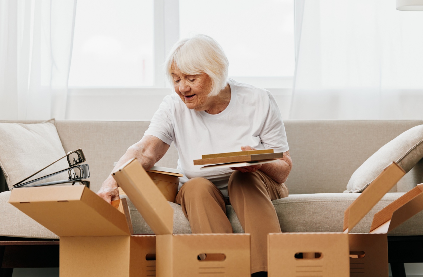 A smiling senior woman sitting on a sofa downsizing her home and sorting belongings into 3 boxes.