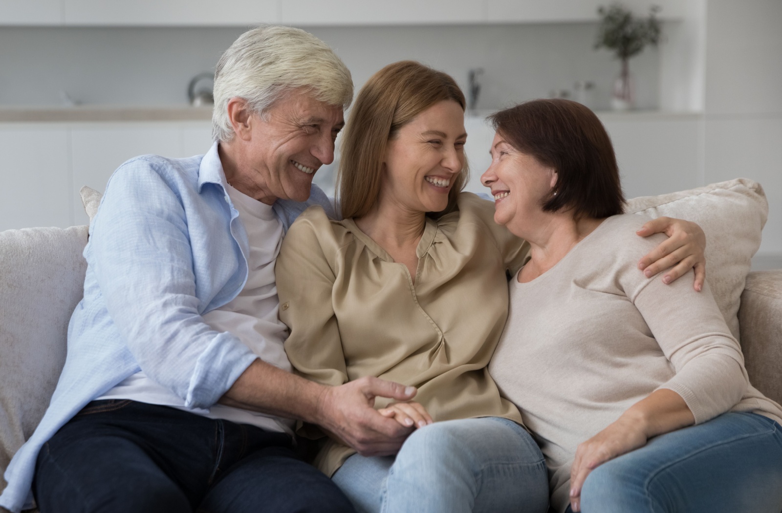 An adult woman sitting between her senior parents on the couch laughing while discussing assisted living.
