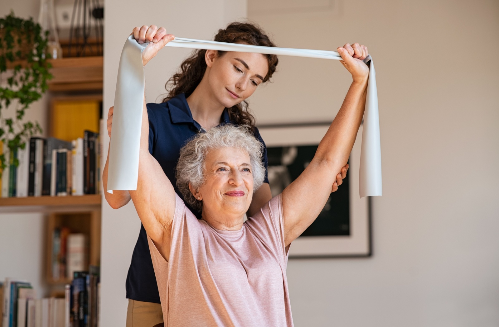 An older adult in senior living lifting her arms overhead with a resistance band while a caregiver supports her.
