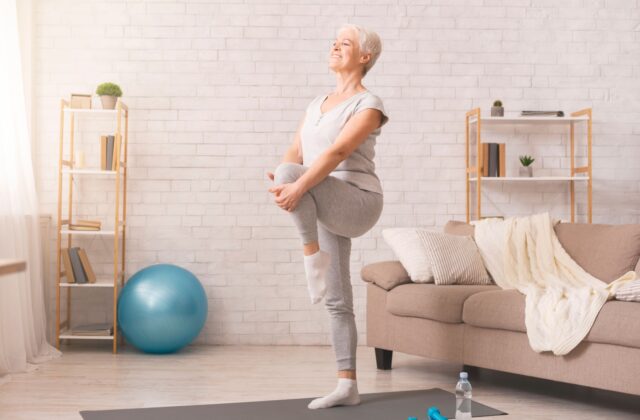 A senior in a sunlit living room lifting their left knee with their hands during a leg-strengthening exercise.