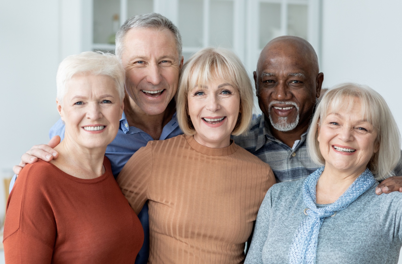 A group of 5 older adults in senior living smiling with their arms around one another.
