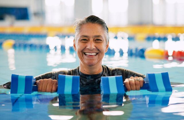 A senior smiling in an indoor pool holding 2 large foam pool weights during water aerobics.