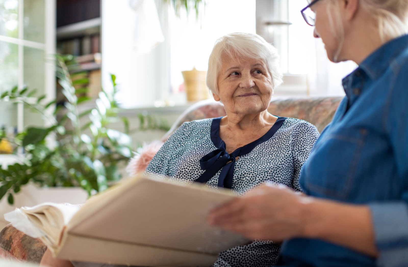 An elderly woman smiling warmly as she shares a moment with a caregiver, looking through a photo album in a cozy, sunlit room.